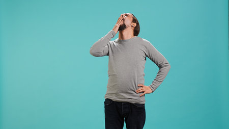 Man feeling exhausted and yawning, covering his mouth, isolated over studio background. Person being...