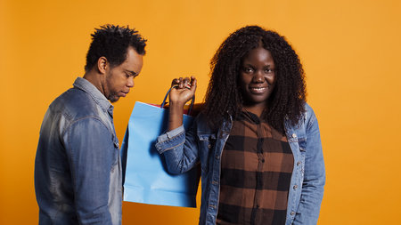 Anxious african american husband getting worried about spending money after his wife purchased many clothes in shopping bags. Young woman feels pleased with buying things on sale. Camera B.の写真素材