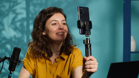 Smiling woman in studio using smartphone on selfie stick to record video, greeting subscribers....