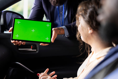 Car salesman showing product offerings on green screen tablet to woman, making recommendations. Showroom agent presents financing options to customer during test drive using chroma key deviceの写真素材