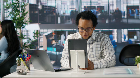 Black man employee joins telecommuting session at office desk, multitasking between project management and business strategy discussions. Productivity and collaboration across departments. Camera A.の写真素材