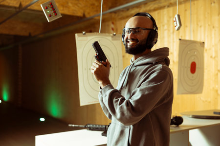 Portrait of happy Arab man in shooting gallery doing training, learning firearm safety and proper gun handling. Smiling Middle Eastern person practicing with pistol, wearing safety glassesの写真素材