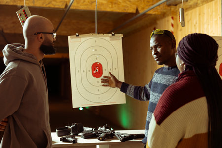 Shooting range safety officer ensuring clients are following rules and operating firearms safely, observing gun holding etiquette. African american firing range gallery worker talking with customersの写真素材