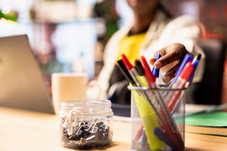 Close up of student in home office reaching for pen from desk organizer, preparing to jot down important notes. Focus on workspace stationary holder used by person in blurry background to do writingの写真素材