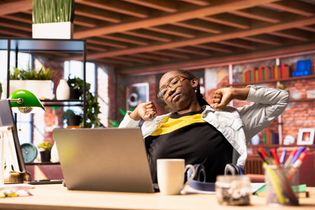 Remote worker in apartment stretching joints after sitting at desk too long. African american woman in home office doing muscle exercises after feeling pain from incorrect postureの写真素材
