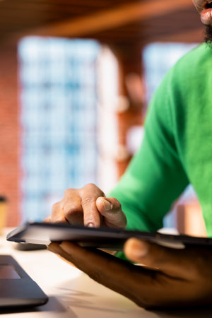 Man in living room using tablet to compose social media posts, producing content. Close up of african american person monitoring social media engagement using digital deviceの写真素材