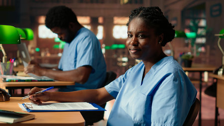 African American nurse in training reviews anatomy case files in a vintage library. Med school student in scrubs studying from database books and research papers in the campus. Camera B.の写真素材