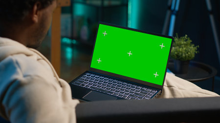 African american person using device with green screen from the comfort of his modern trendy home, sitting on the couch and working in the living room. Happy confident young man. Camera A.の写真素材