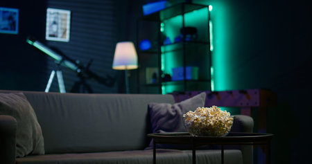 Apartment room with dimmed lighting, popcorn and remote on small table, creating relaxed mood for movie night, close up. Panning shot of popcorn bowl on table in living roomの写真素材