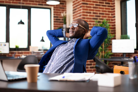 Black businessman leaned on office chair with hands behind his head, relaxing after busy schedule in startup company. Male employee at desk, stretching his back and feeling satisfied after work.の写真素材
