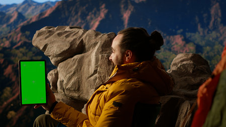 Adventure seeker stands at a mountain peak next to isolated green screen, looking out at the scenic...