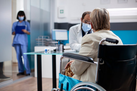 Senior woman in wheelchair sharing her medical concerns with young doctor during clinic visit. Female pensioner patient receiving detailed advice about her health from male physician in hospital room.の写真素材