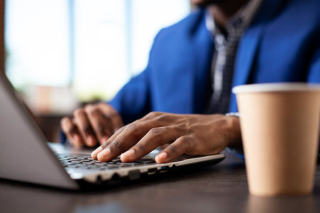 African american person typing on digital device at desk in modern workspace. Selective focus on hands inputting information for business project on laptop with coffee cup placed nearby on table.の写真素材