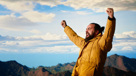 Satisfied adventure seeker stands at the top a towering mountain, taking in the epic view and feeling proud of his hiking achievement. Strong joyful trekker feels the rewarding experience. Camera B.の写真素材