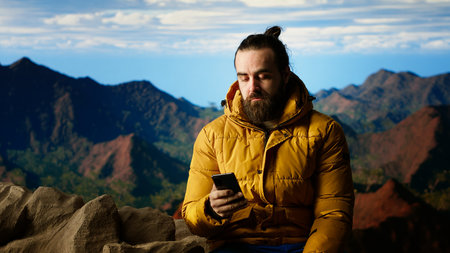 Nature lover relaxing on mountain top getting online with his phone at a scenic viewpoint, looking out over the landscape. Hiker staying always online and connected, panoramic scenery.の写真素材
