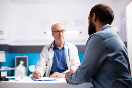 Elderly healthcare specialist in medical office talks with male patient, taking notes on his clipboard. Professional old physician listening to health concerns of young man during clinic appointment.の写真素材