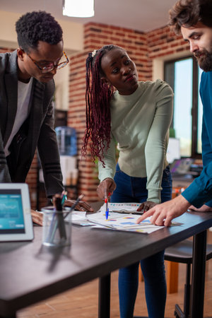 African american female manager with pen in hand, speaking to male employees in brick wall office. Businesswoman points at documents, having company project discussion with colleagues at table.の写真素材