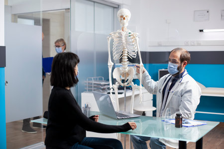 Caucasian doctor explains spinal recovery options to female patient in clinic room, using skeletal model for visual aid. Physician points at skeleton and discusses bone treatment plan with sick woman.の写真素材