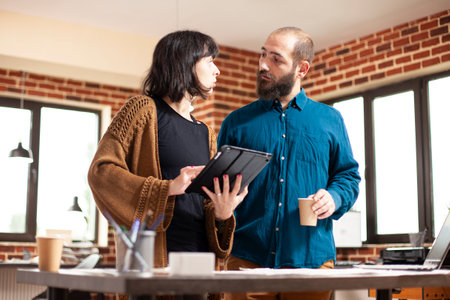 Businesswoman holding tablet and listening to male colleague, sharing strategy ideas for startup company. Bearded man and coworker having project development discussion in modern brick wall office.の写真素材