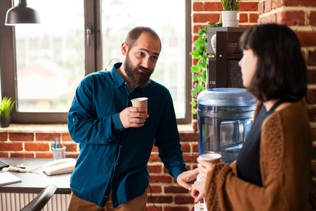 Businessman and woman share conversation near water cooler in brick wall workspace. Male and female employees hold coffee cups combining casual interaction with productive talk in startup office.の写真素材