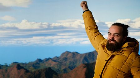 Triumphant hiker stands at the mountain peak with his arms raised in victory, celebrating success and personal achievement of climbing another mountain. Breathtaking panoramic view. Camera B.の写真素材