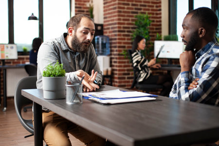 Caucasian man seated at desk, describing his work experiences to african american recruiter. Black hr manager listening to bearded male candidate during job interview in brick wall office.の写真素材