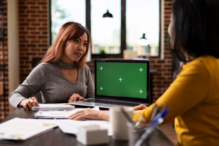 Laptop displaying greenscreen as asian businesswoman speaks to her coworker at desk, discussing marketing strategies. Female colleagues share project plans, using digital device with mockup template.の写真素材