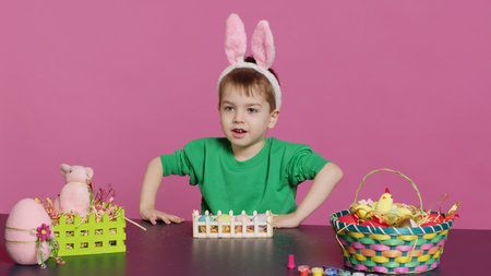 Happy toddler with bunny ears arranging basket filled with painted eggs, creating festive...