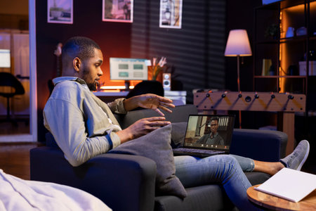 Focused black guy smiling and waving during a web conference in his living room, showcasing online...
