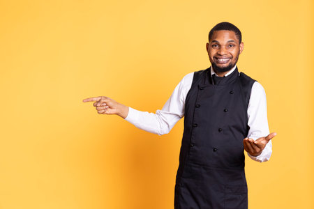 Smiling friendly butler pointing direction to left or right sides against yellow background, creating an advertisement for the catering industry. Five star restaurant employee.の写真素材