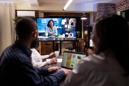 Multiethnic colleagues analyzing reports in late night meeting. Digital screen shows a videoconference call in progress, highlighting remote collaboration and business strategy in modern workspace.の写真素材