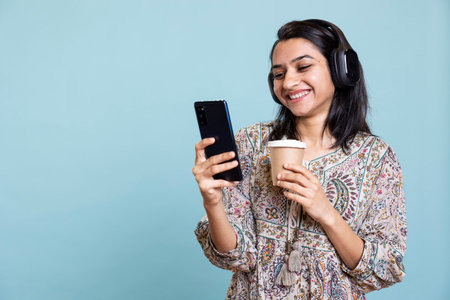 Pleased excited woman playing music on phone and drinking coffee, having fun dancing on her favorite...