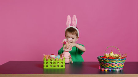 Joyful young kid playing around with festive painted decorations, showing a rabbit toy and a pink...
