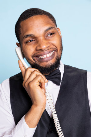 African american hotel porter provides crucial lodging information via landline phone call, discussing about a room reservation with guests. Bellhop answering questions remotely.の写真素材