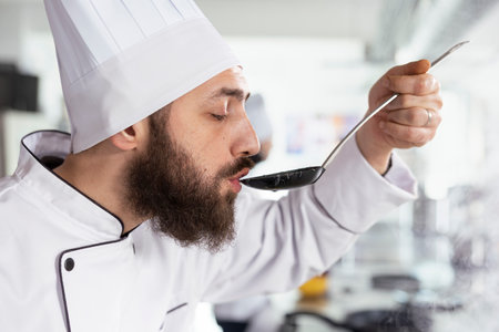 Focused on culinary perfection, male head chef in uniform uses a spoon to taste a delicious soup, adjusting the seasoning for a gourmet dish. Kitchen filled with aromatic ingredients. Close up.の写真素材