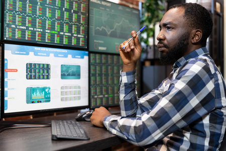 Focused black man working late at a desk, analyzing stock market data on multiple monitors in a modern office. Stockbroker holding a pen as he contemplates and reviews trading statistics.の写真素材