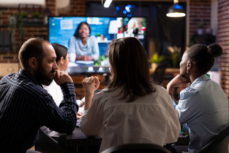 Business professionals collaborate in nighttime office meeting. A videoconference call is displayed on digital monitor as team members discuss marketing strategies and review financial data.の写真素材