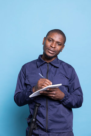 Security agent in safeguarding uniform takes notes on a notebook, standing against blue background. Serious dedicated bodyguard writing info to keep track of safety responsibilities.の写真素材