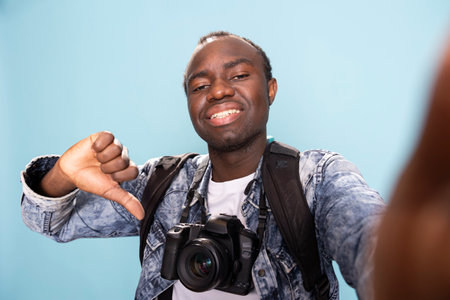 Happy african american photographer with a professional camera around his neck, giving thumbs down in studio. POV of a black male vlogger recording himself while making a playful disagreeing gesture.の写真素材