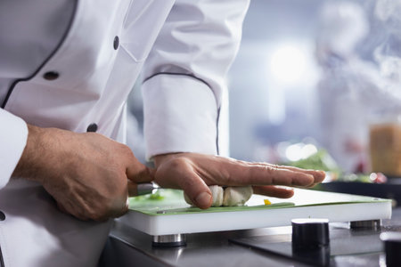 Young chef with hat making crushed garlic with knife on the cutting board, pressing cloves to use condiments for gourmet dish in the restaurant kitchen. Male cook uses spices at counter. Close up.の写真素材