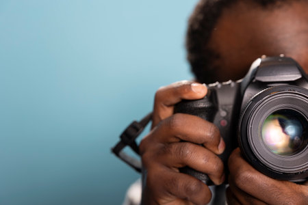Closeup of black male photographer using a professional camera in a studio with a blue background. Selective focus on an african american man holding and demonstrating how to use a DSLR.の写真素材
