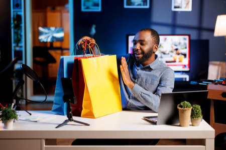 Man filming fashion vlog in apartment, holding shopping bags in hand, showing subscribers purchases he recently got. Media star presents viewers new acquisitions, filming himself at homeの写真素材