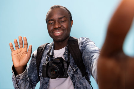 African American man with DSLR around his neck, makes hello gesture, recording a video for his vlogging channel. Black male photographer smiles and waves at the camera in studio with blue background.の写真素材