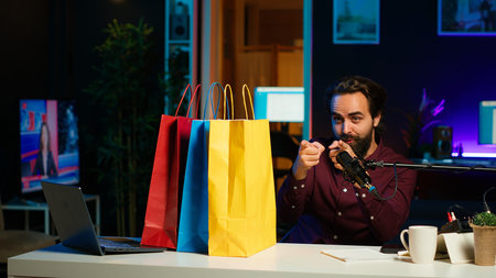 Excited media star holding colorful shopping bags, showing fans purchases, filming himself in apartment. Playful content creator in studio presenting audience new acquisitions, camera Aの写真素材