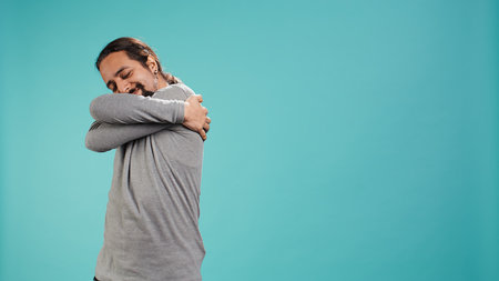 Portrait of man standing in studio, hugging himself in gesture of comfort and self love, isolated over studio background. Cheerful person doing self acceptance, embracing himself, camera Bの写真素材