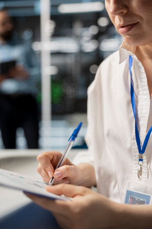 Woman in data center taking note on fixes to be made after running diagnostic scripts on computer. Server room admin writing repairing needed by supercomputers after evaluating performance metricsの写真素材