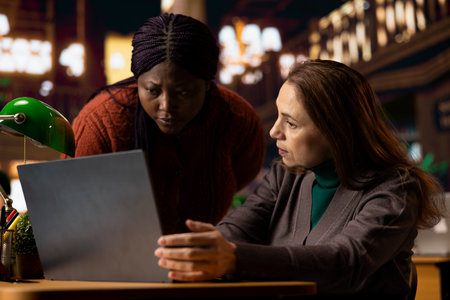 Mature students at a library desk studying materials on a laptop, reading and collecting information from web archive files. Diverse team of pupils sharing notes, research for degree.の写真素材
