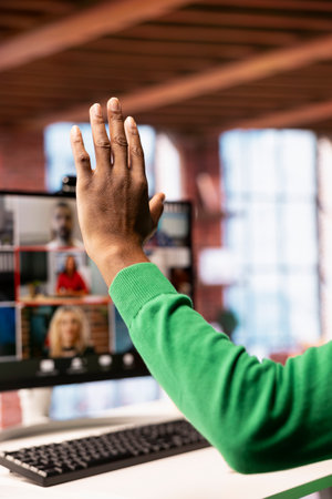 Teleworker at home office desk raising hand to request turn to speak during online videocall on PC....