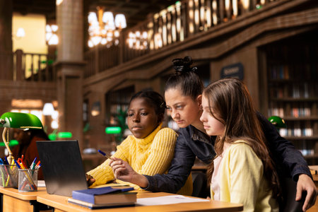 Tutor guiding two elementary school girls at a library desk, providing lessons to aid in their...