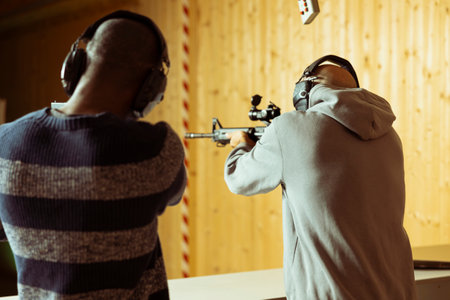 Friends having fun in shooting gallery using rifles to hit targets. Mates entertained by shooting bullseyes at indoor firing range facility, competing against each otherの写真素材